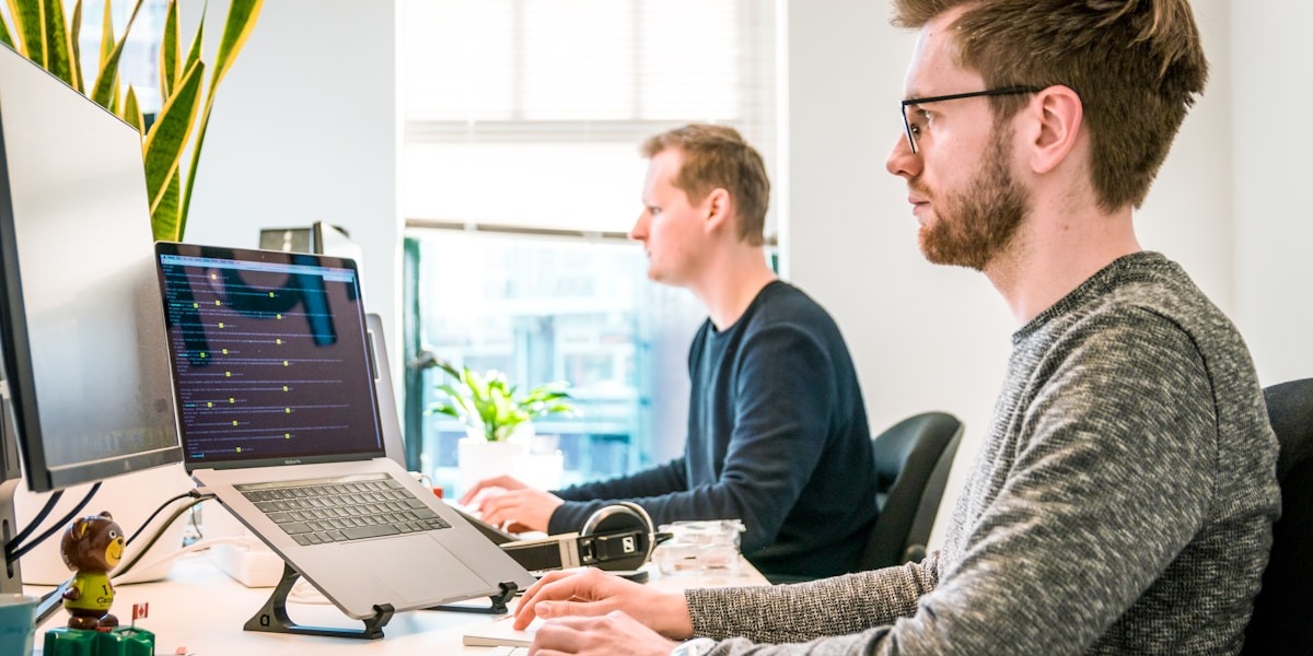 Developers working at computers in an office, monitoring code and system activity on multiple screens.