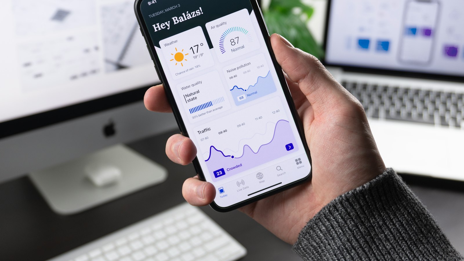 Hand holding a smartphone showing a data dashboard with traffic and air-quality charts, in front of two desk monitors
