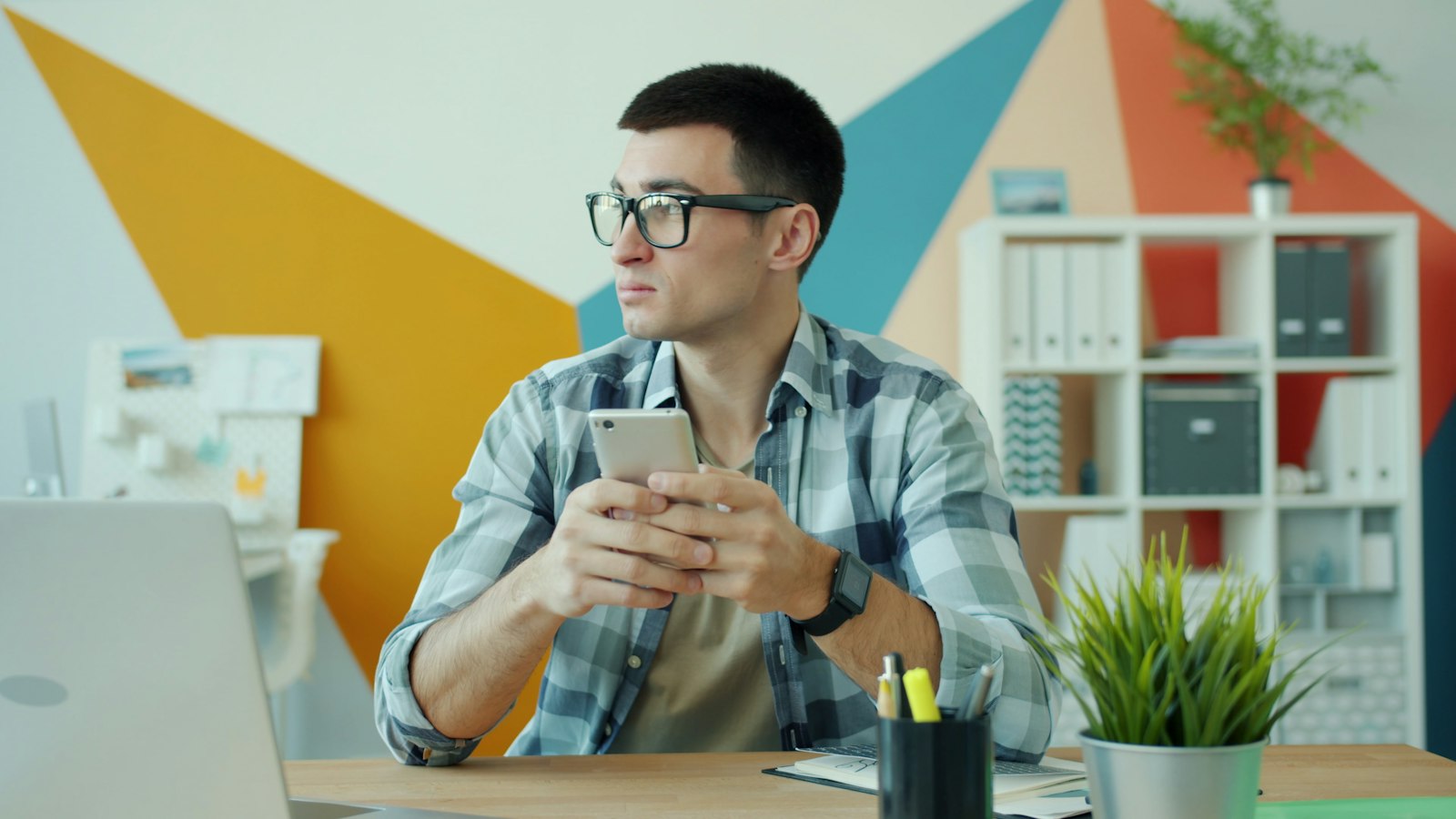 Man in a checked shirt and glasses using a smartphone at an office desk with a laptop, a plant and a colourful wall behind him